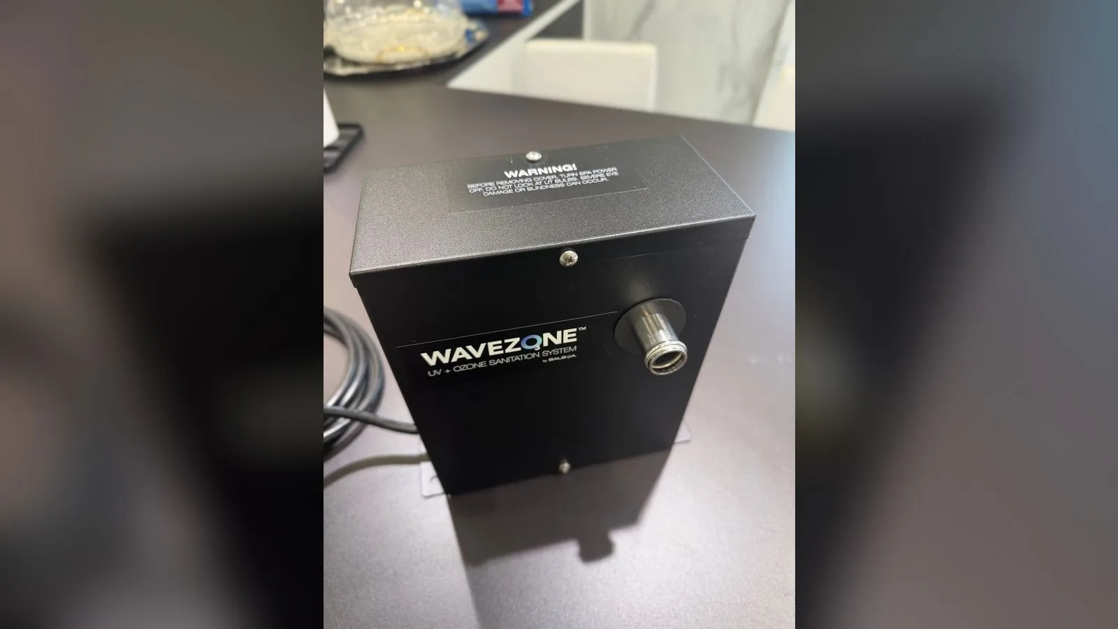 Boîtier noir Balboa Wavezone UV + Ozone Sanitation System, avec son étiquette d'avertissement sur le capot et son raccord chromé en façade