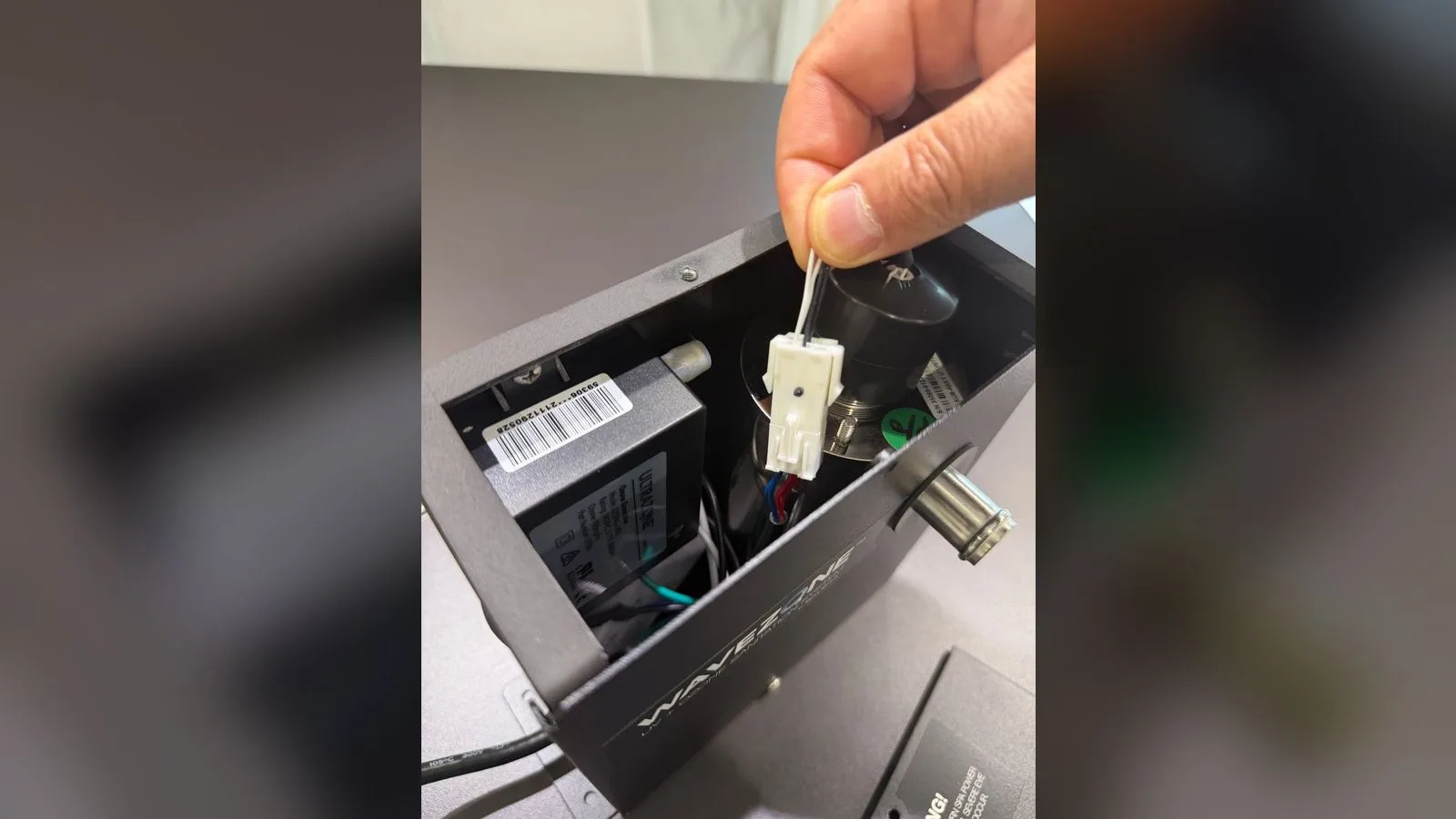 Doigts tenant la prise blanche du câble de la lampe UV, encore branchée, à l'intérieur du boîtier Wavezone